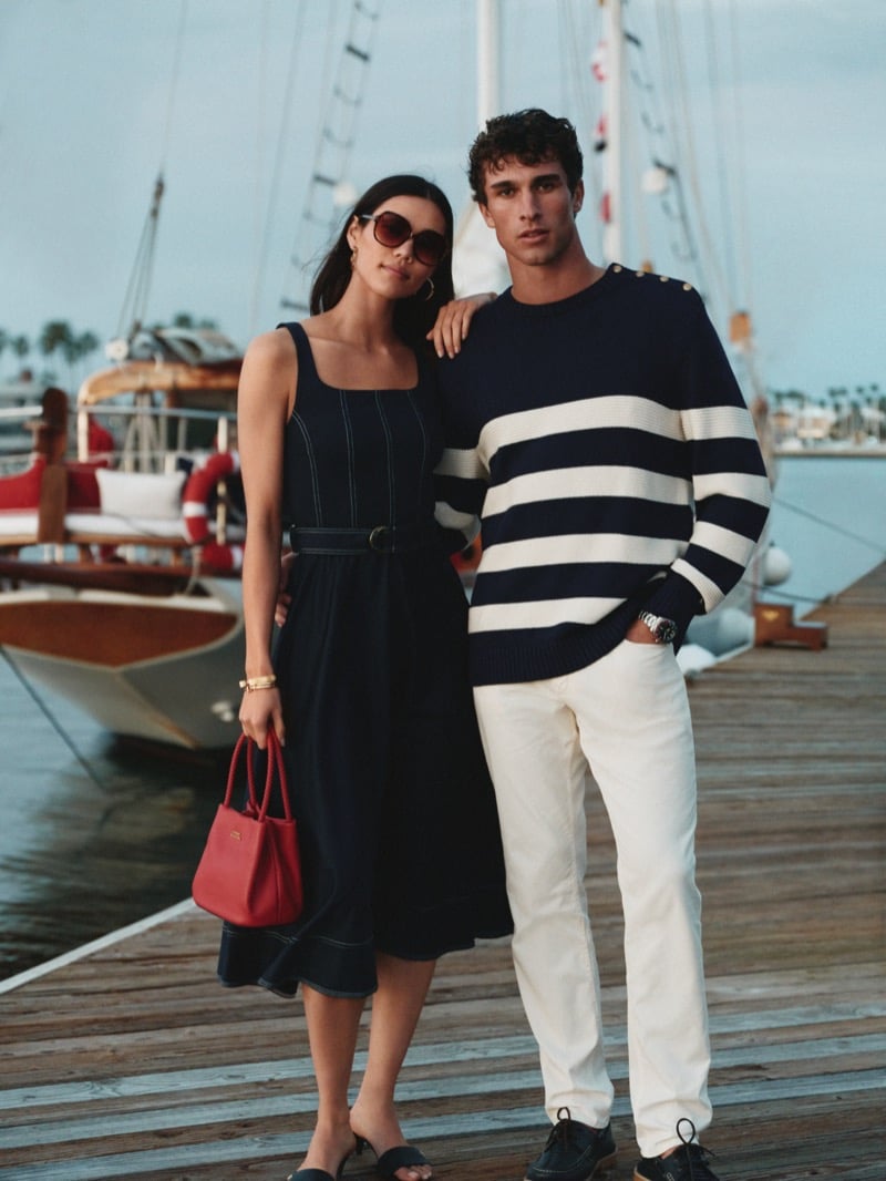 Model Kai Paula stands shoulder to shoulder with a female cast member on a marina dock for Tommy Hilfiger summer 2026, wearing a striped crewneck sweater with shoulder buttons over light trousers, beside a moored sailboat