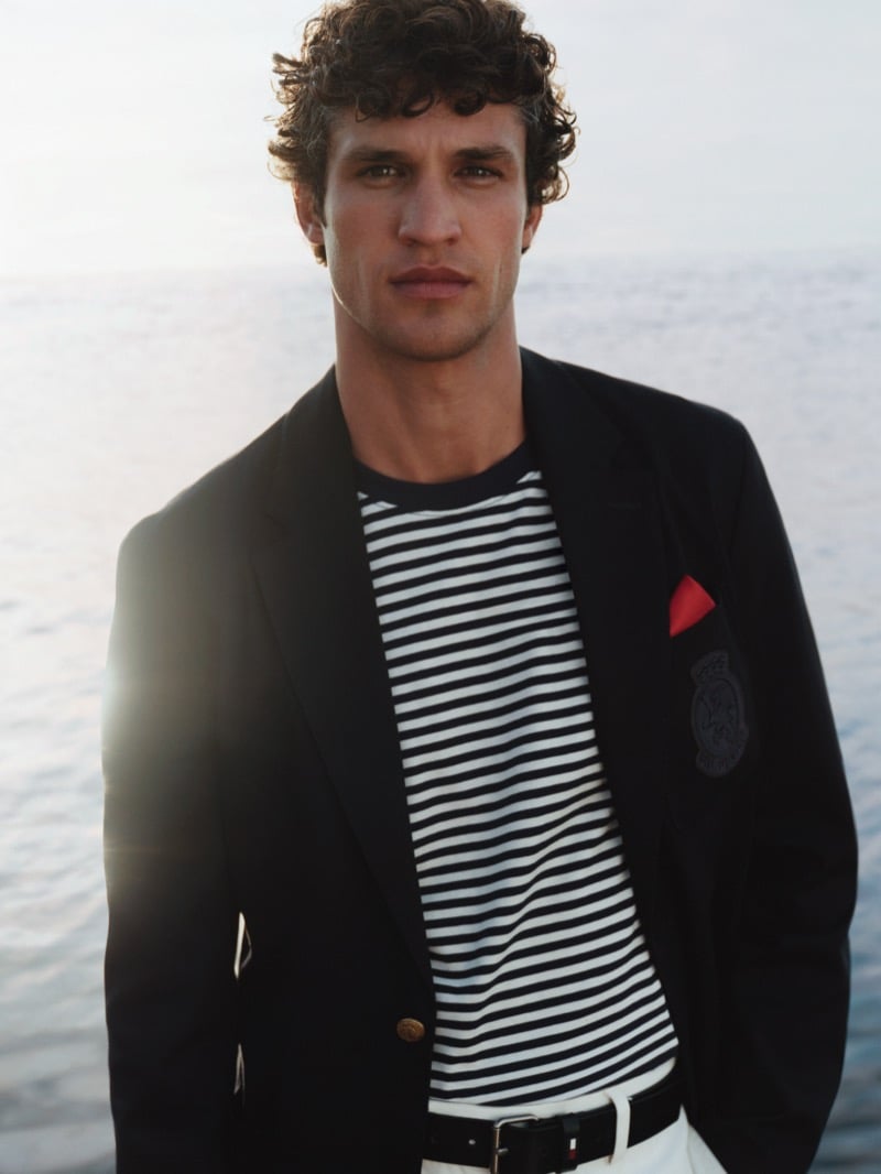 Model Francisco Henriques wears a Tommy Hilfiger summer 2026 dark blazer with a crest patch over a Breton-stripe crewneck tee, finished with a red pocket square and white trousers, set against an out-of-focus sea