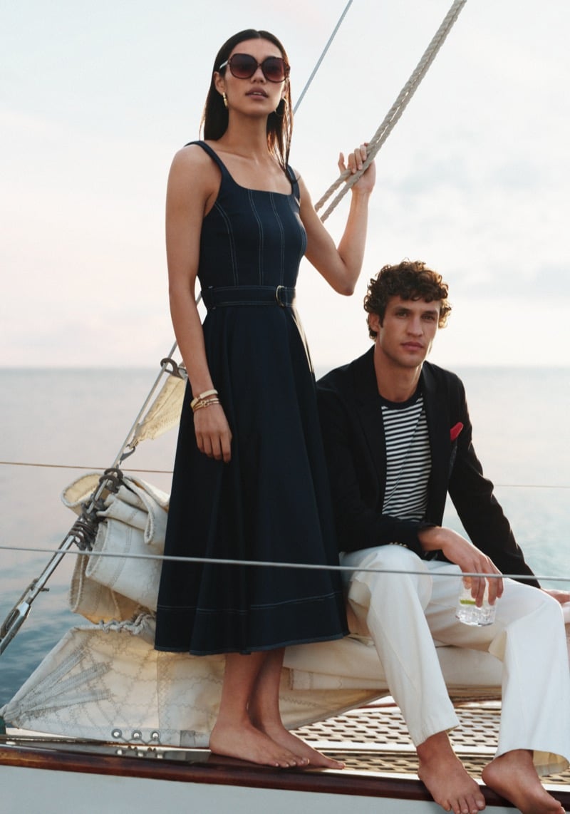Francisco Henriques sits on the bow of a sailboat in a Tommy Hilfiger summer 2026 dark blazer and Breton-stripe tee with light trousers, beside a female model in a belted sleeveless midi dress holding a rigging rope