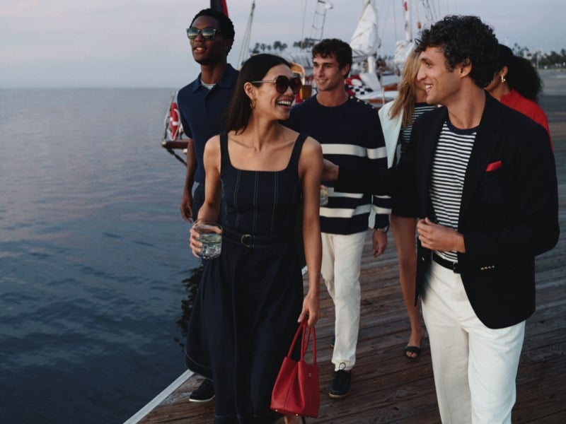 Tommy Hilfiger summer 2026 dockside group shot with Hamid Onifade in a tipped polo, Kai Paula in a striped marine pullover, and Francisco Henriques in a blazer over a Breton tee, walking alongside female cast members at sunset