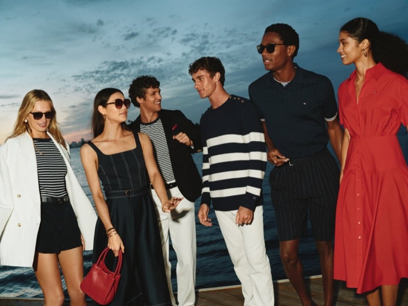 Tommy Hilfiger summer 2026 cast portrait at the marina with Francisco Henriques in a crested blazer over a Breton tee, Kai Paula in a striped crewneck, and Hamid Onifade in a tipped piqué polo alongside female cast members in dresses, knits, and tailored shorts