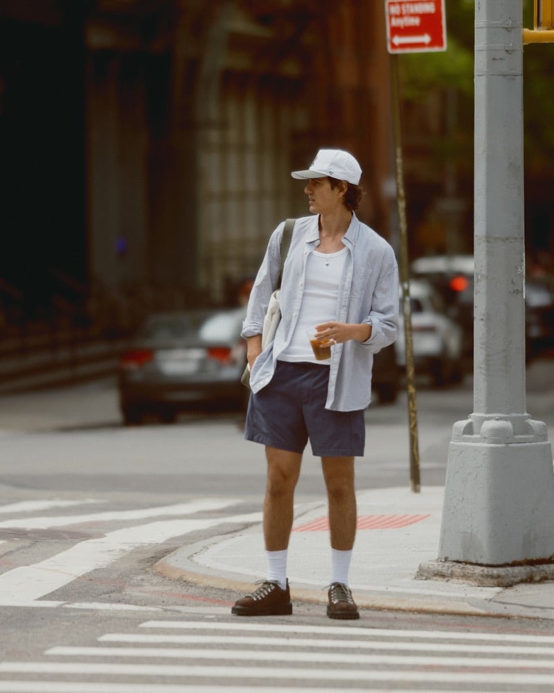 Model Tony Özkan stands at a downtown New York crosswalk in a white 47 baseball cap, a blue and white striped button-up shirt open over a ribbed tank, navy pull-on shorts, a canvas tote, white crew socks, and brown suede hiking-inspired shoes, holding an iced coffee for Abercrombie & Fitch