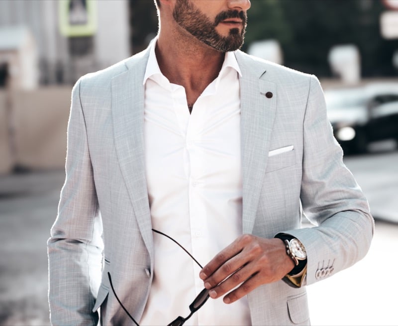 Bearded man in a pale gray tropical wool suit jacket with notch lapels and a small pocket square, holding sunglasses in one hand with a wristwatch at the cuff, an urban background blurred behind him