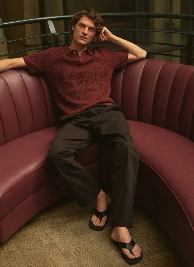 Valentin Caron reclines on a curved burgundy banquette in a deep red knit polo and dark wide-leg trousers, paired with dark thong sandals