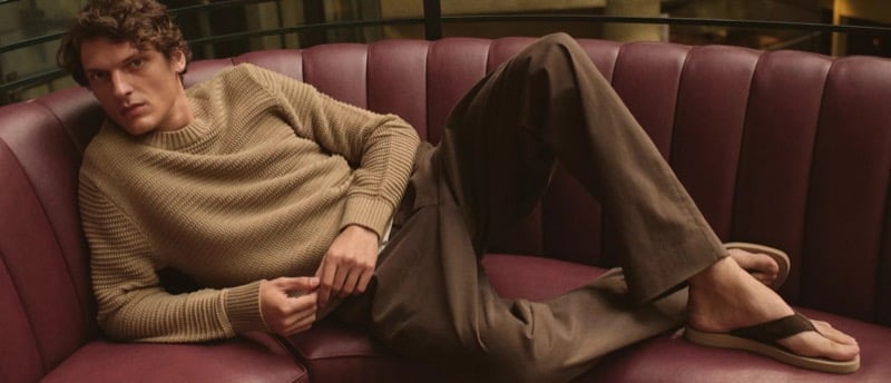 Valentin Caron reclines on a curved burgundy banquette in a camel ribbed crewneck sweater, brown trousers, and brown thong sandals