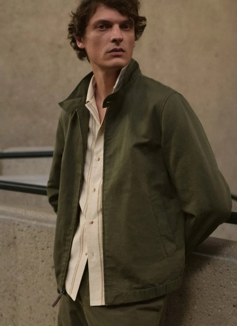 Valentin Caron stands on a concrete landing in an olive cotton harrington jacket layered over a cream striped camp-collar shirt with matching olive trousers