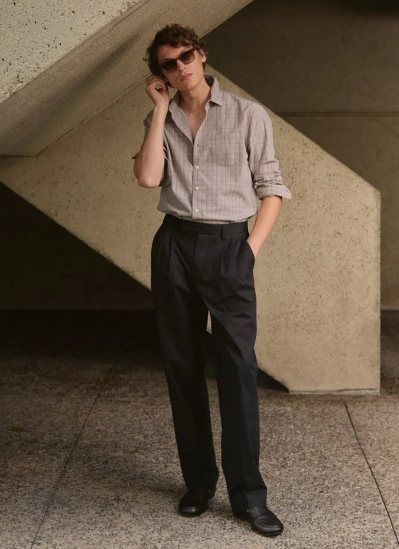 Valentin Caron stands beneath a concrete pylon in tortoise sunglasses, a relaxed micro-check button-down with rolled sleeves, dark wide-leg pleated trousers, and dark loafers