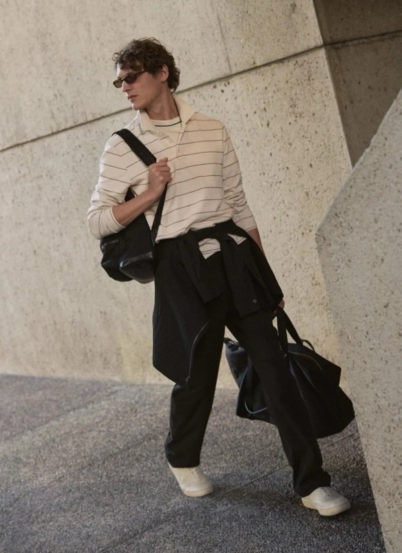 Valentin Caron walks past a concrete wall carrying a soft leather backpack and a tote, wearing a cream long-sleeve striped polo, a jacket tied around his waist, and trousers with cream sneakers