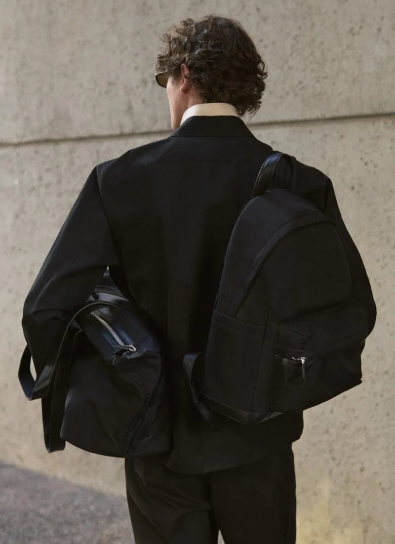 Valentin Caron walks away from the camera, carrying a soft tote bag and an oversized backpack