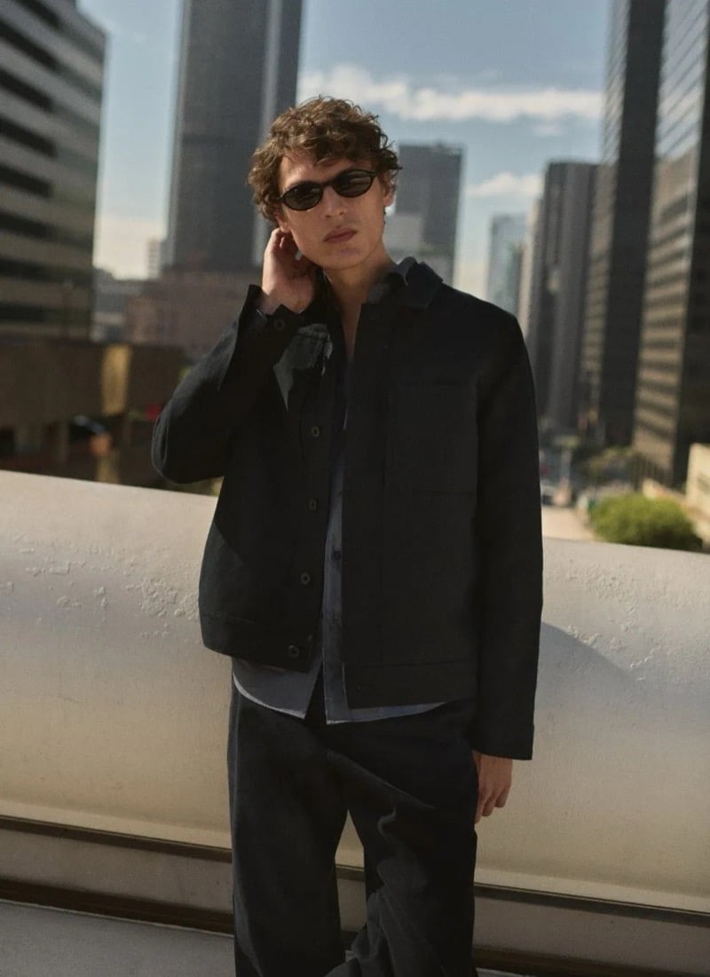 Valentin Caron stands on a downtown Los Angeles terrace in a dark chore jacket over a tonal shirt and pleated trousers, with a high-rise skyline behind him