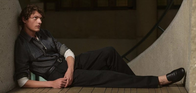 Valentin Caron reclines on a tiled floor in a chambray shirt with rolled sleeves, a slim leather belt, dark pleated trousers, and dark leather loafers