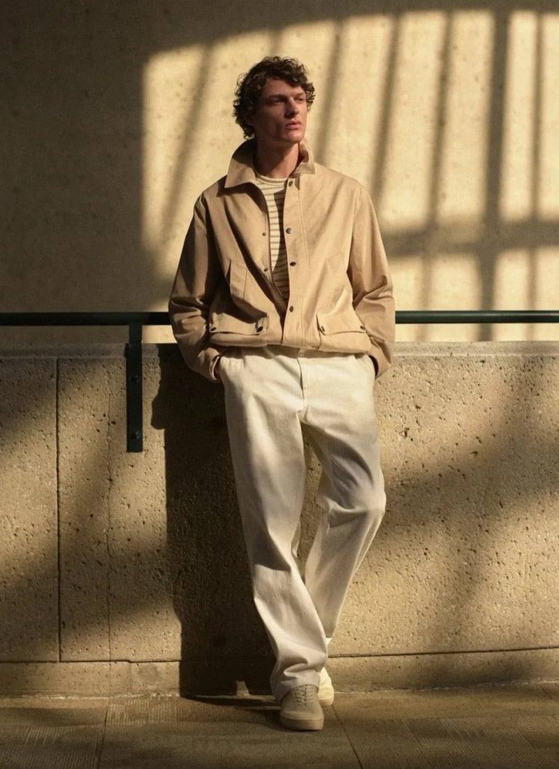 Valentin Caron leans against a concrete wall in a tan cotton harrington jacket over a striped knit tee and relaxed cream trousers with cream canvas sneakers