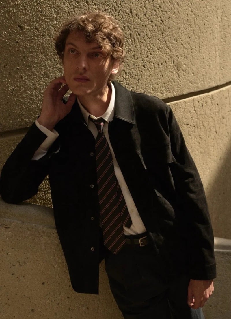 Valentin Caron in a dark chore jacket layered over a light shirt and a striped tie with a slim leather belt, set against a curved concrete wall