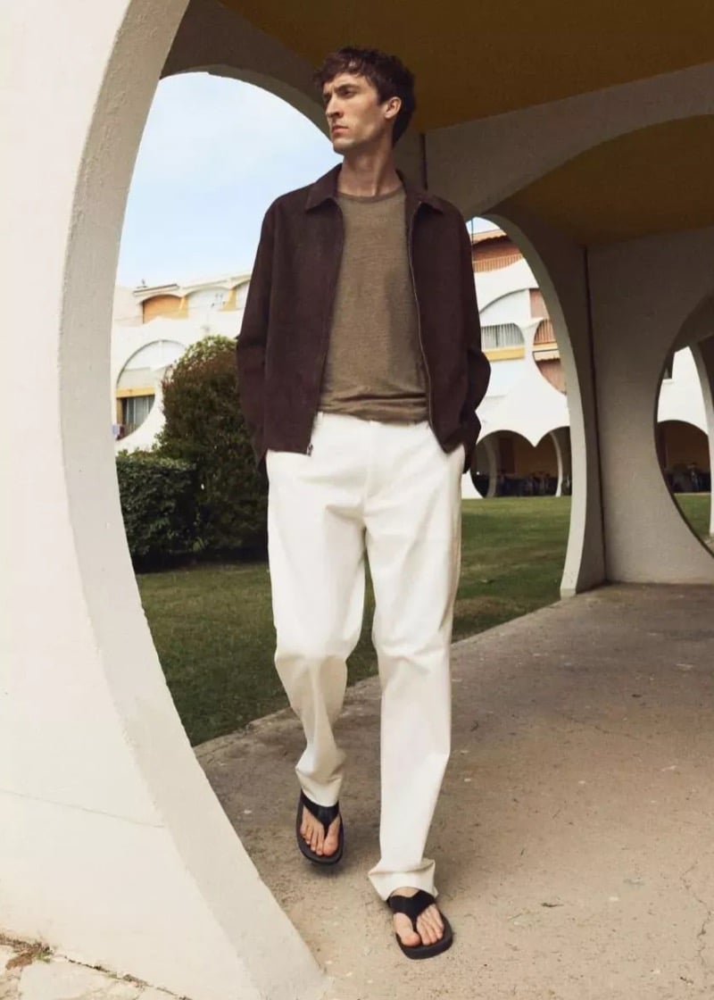 Tim Dibble stands framed by a large circular archway in a brown zip harrington jacket over a taupe lightweight knit, off-white straight-leg trousers, and dark flip-flops