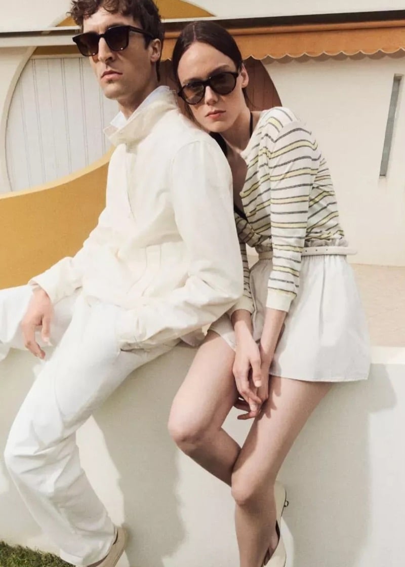 Tim Dibble and a female model perch on a low wall in matching pale-toned looks, Tim in a sporty jacket and pants, his companion in a striped knit belted over a full skirt, both in sunglasses