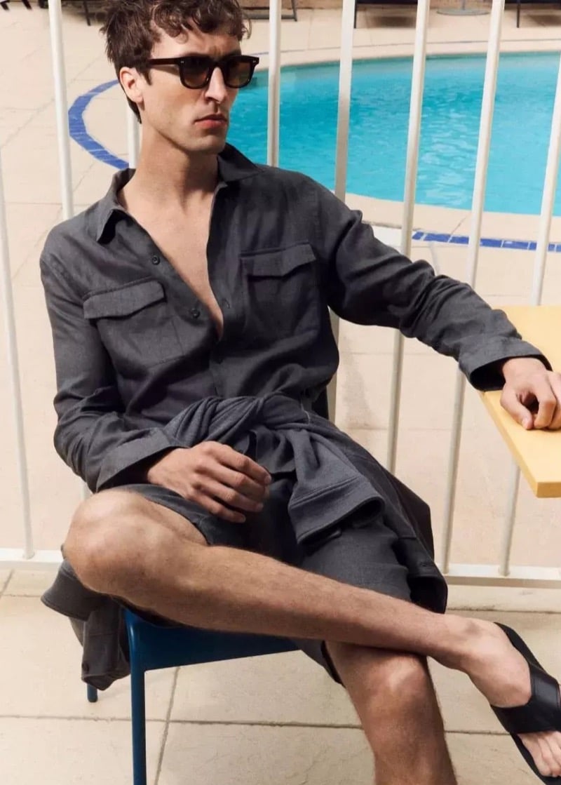 Tim Dibble sits poolside on a blue chair in a charcoal utility shirt with chest patch pockets, matching shorts, sunglasses, and leather sandals