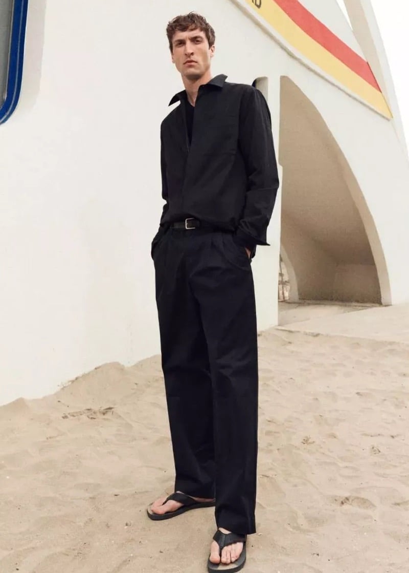 Tim Dibble stands on the sand in front of a modernist lifeguard tower in a black shirt and pleated trousers with a leather belt, and leather flip-flops
