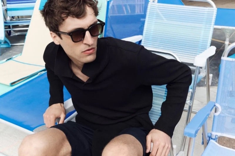 Tim Dibble sits in a pale blue vintage folding beach lounger in a black long-sleeve knit polo, shorts, and sunglasses