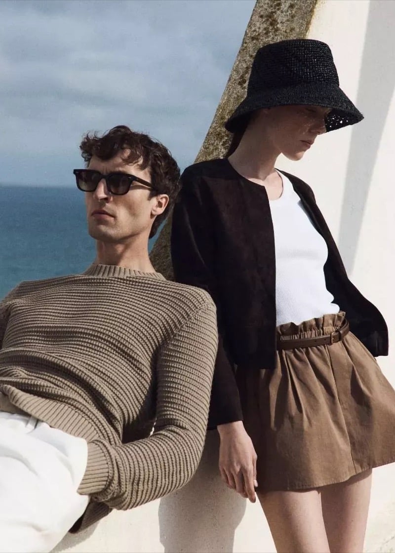 Tim Dibble leans against a concrete wall by the sea in a camel ribbed crewneck sweater, joined by a female model in a dark cardigan, ribbed tank, tan paperbag skirt, and dark straw bucket hat