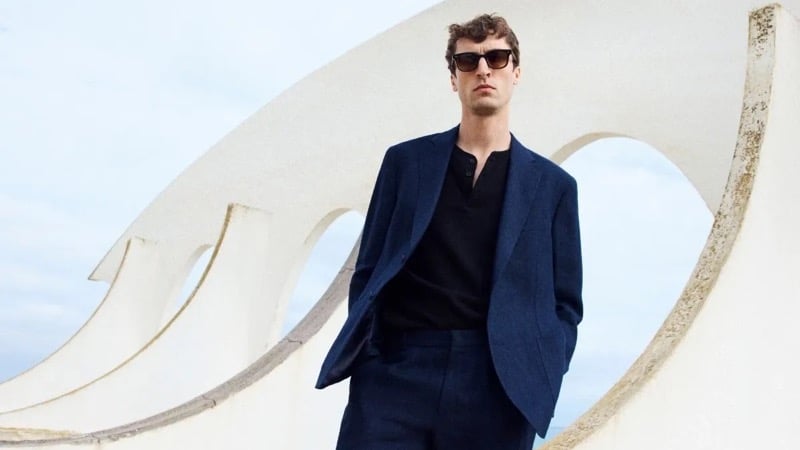 Tim Dibble stands in front of a sweeping sculptural concrete arch under an open sky, wearing a navy tailored blazer over a black henley with navy trousers and sunglasses