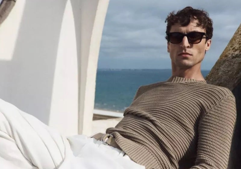 Tim Dibble reclines against a concrete arch overlooking the Mediterranean in a camel ribbed crewneck sweater and tortoise sunglasses