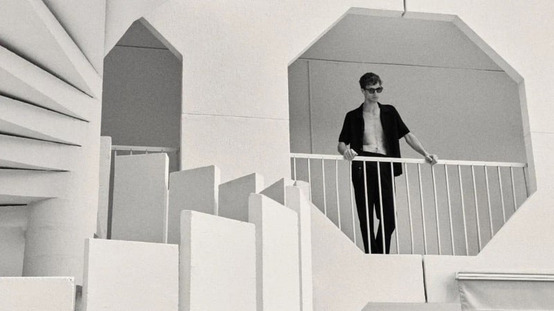 Black-and-white image of Tim Dibble standing behind a railing beneath a huge octagonal window cutout in a modernist concrete building, wearing an open short-sleeve shirt over dark trousers