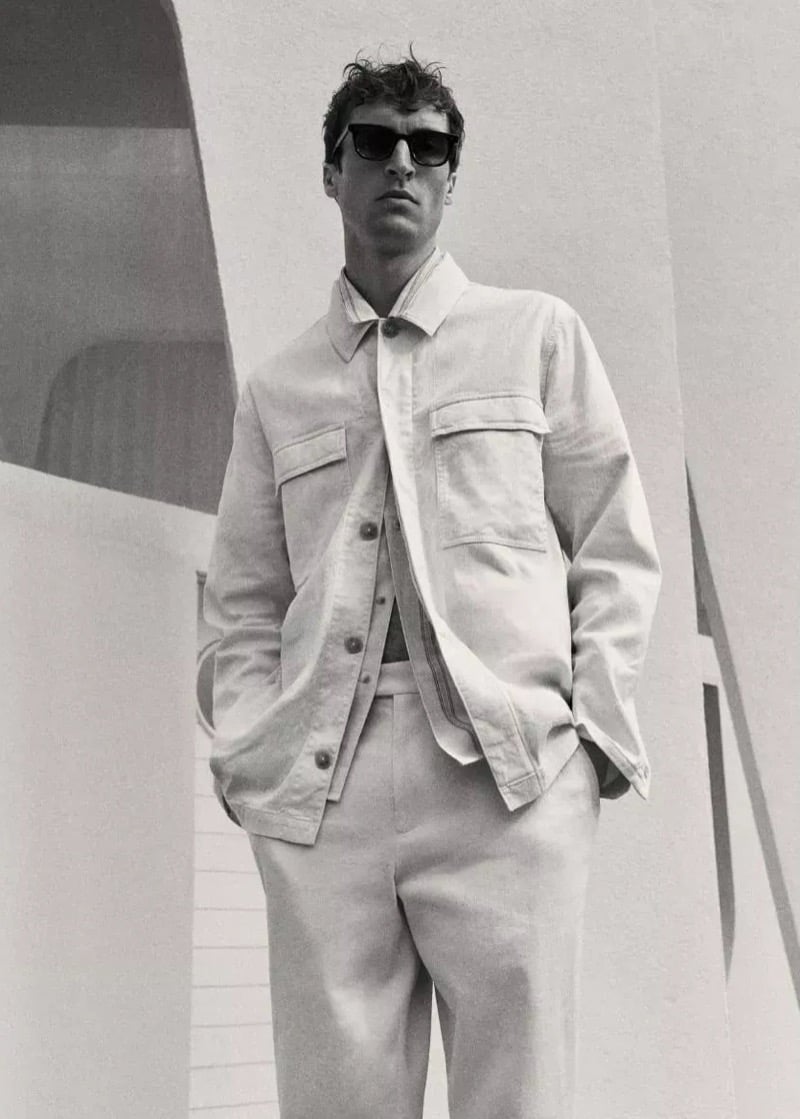 Black-and-white portrait of Tim Dibble in a boxy overshirt layered over a shirt, paired with straight trousers, standing before sculptural modernist forms