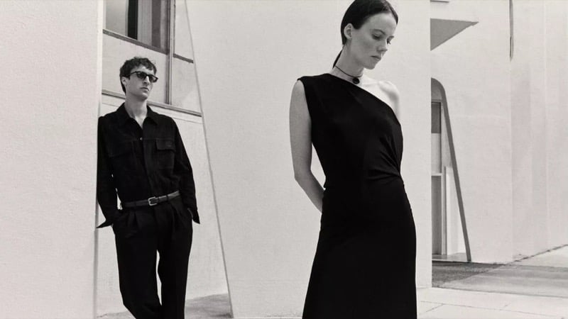 Black-and-white image of Tim Dibble leaning against a sculptural wall in a dark utility shirt, belt, and pleated trousers while a female model in a one-shoulder dark dress stands in the foreground