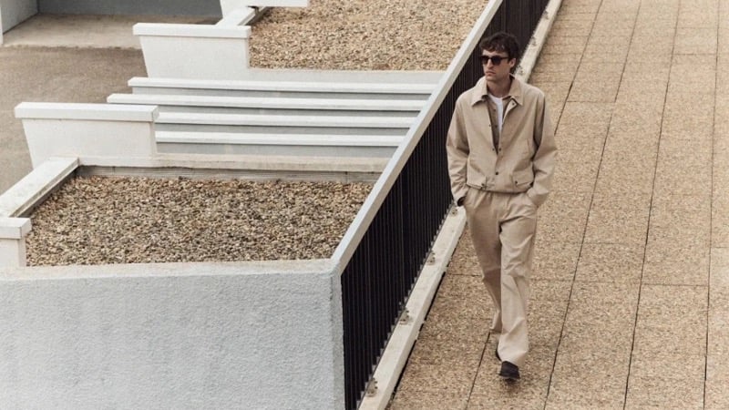 Tim Dibble walks along a stepped boardwalk beside a gravel-filled architectural planter in a sand-toned zip jacket and matching pants