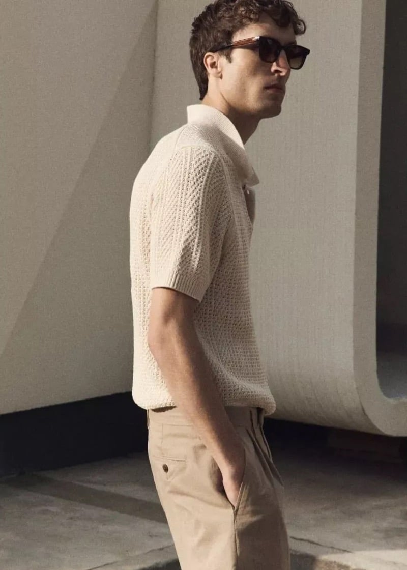 Tim Dibble stands in profile in an ecru open-knit crochet polo and tan trousers, framed by sculptural concrete forms