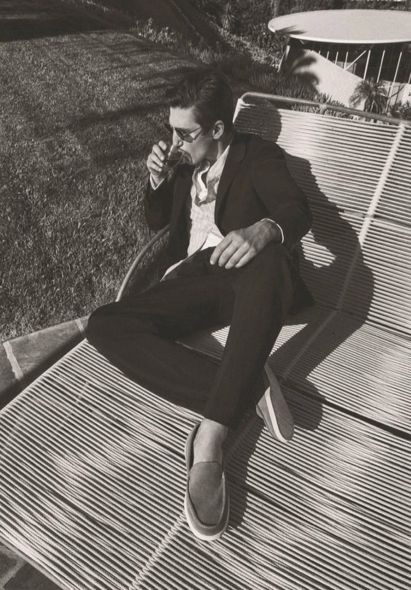 Black-and-white image of Anatol Modzelewski lounging on a rope chaise in a dark tailored suit and suede loafers, sipping from a glass, for Vince spring 2026