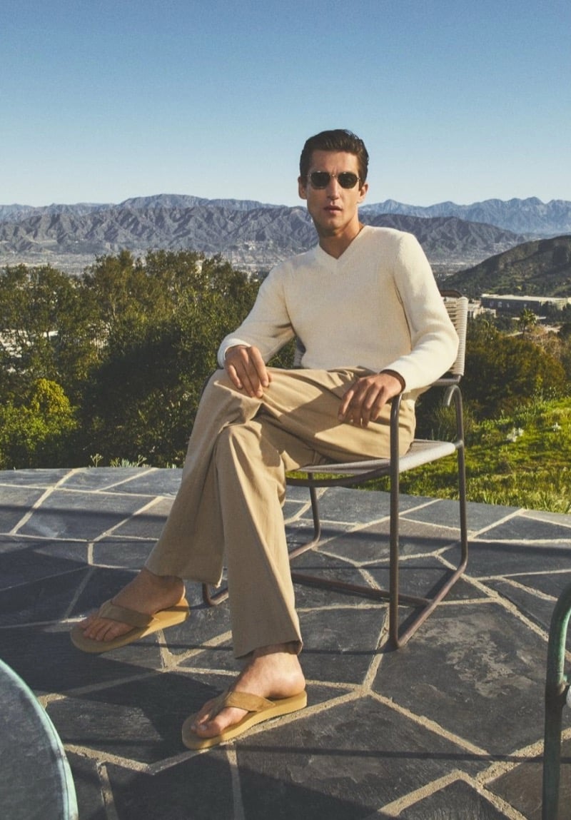 Anatol Modzelewski wears a cream V-neck sweater with wide-leg khaki trousers and tan suede sandals seated on a stone terrace overlooking mountains for Vince spring 2026