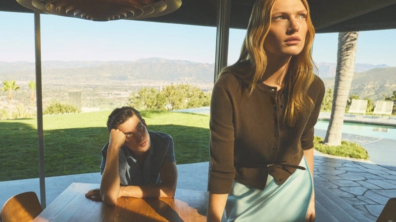 Anatol Modzelewski seated in a slate blue shirt at a wooden table while Liz Kennedy stands in a brown knit cardigan and pale blue skirt inside a modernist Los Angeles home for Vince spring 2026