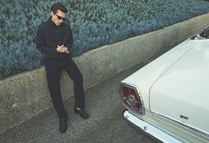 Anatol Modzelewski leans against a retaining wall in a navy zip moto jacket and dark trousers beside a vintage white convertible for Vince spring 2026