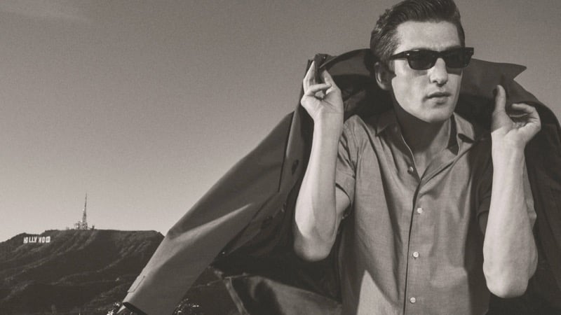 Black-and-white image of Anatol Modzelewski wearing a short-sleeve button-up shirt, sunglasses, and a draped sport coat over his shoulder with the Hollywood sign visible behind him for Vince spring 2026