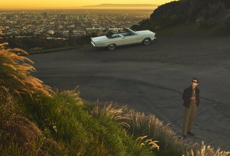 Anatol Modzelewski in a brown jacket and tan trousers stands on a hillside with a vintage white convertible and the Los Angeles skyline at sunset for Vince spring 2026