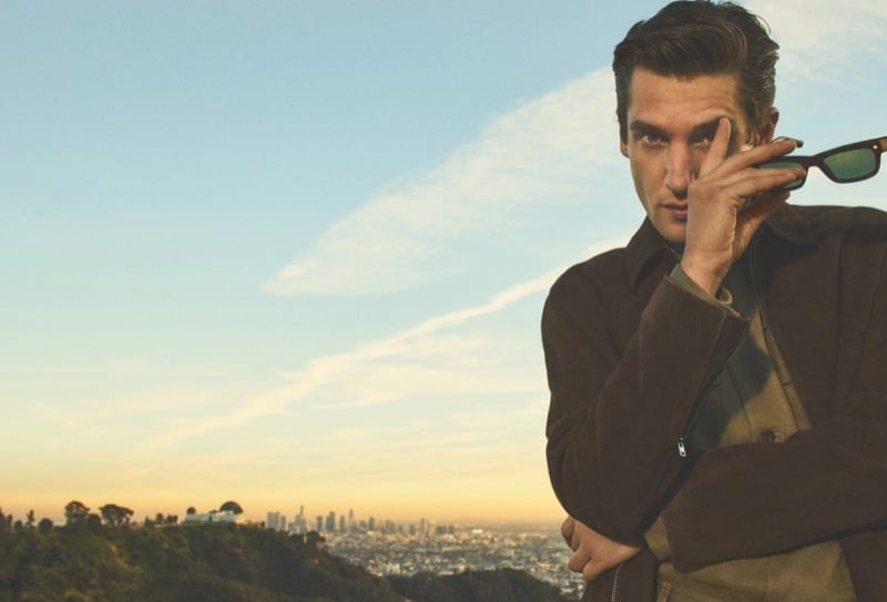 Anatol Modzelewski wears a brown suede jacket over an olive crew neck with sunglasses, photographed above Los Angeles with the city skyline at sunset for Vince spring 2026