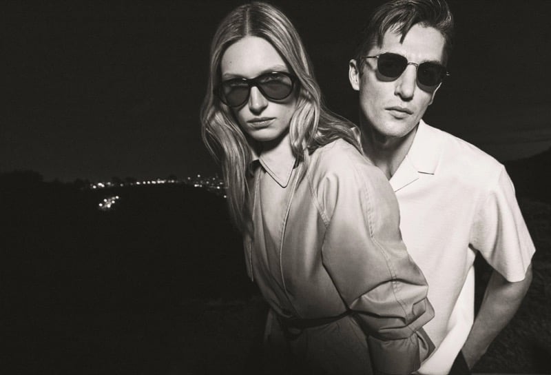 Black-and-white image of Anatol Modzelewski in a short-sleeve shirt and sunglasses standing with Liz Kennedy in sunglasses at night with the Los Angeles lights behind them for Vince spring 2026