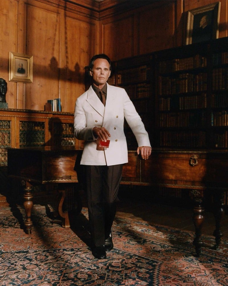 Walton Goggins stands in a wood-paneled library of an English estate in an ivory double-breasted blazer over a dark shirt and tie, holding a small red book at his side
