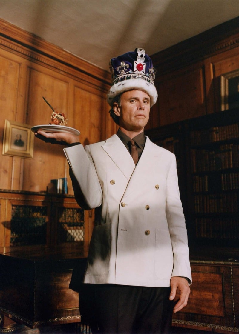 Walton Goggins wears a jeweled royal crown with an ivory double-breasted blazer and dark shirt and tie, balancing a small plate with a single skewered bite on his outstretched hand inside a wood-paneled library