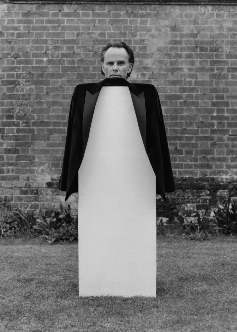 Walton Goggins stands behind a tall plinth on an estate lawn in a peak-lapel evening jacket with his head emerging at the top, photographed in black and white against a brick wall