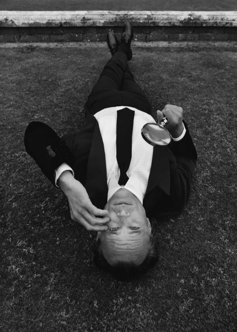 Walton Goggins lies flat on a lawn in evening tailoring with bow tie, holding a magnifying glass up to one eye, in a black and white image shot from above