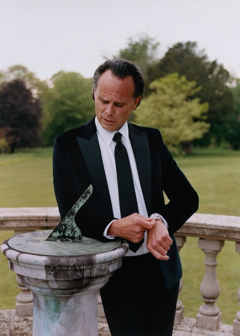 Walton Goggins leans on a weathered stone sundial on a country estate terrace, dressed in a peak-lapel tuxedo and dress shirt with a long tie, checking his watch