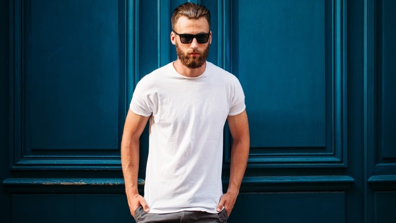 Bearded man in sunglasses wearing a midweight white crewneck tee standing in front of a paneled door, the collar, sleeve hems, and body hem clearly visible for construction inspection