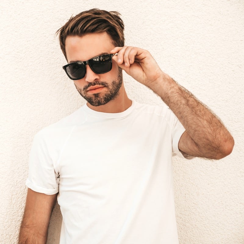 Close portrait of a man in sunglasses wearing a textured white crewneck tee with rolled sleeves against a pale wall, the surface of the cotton knit visible up close