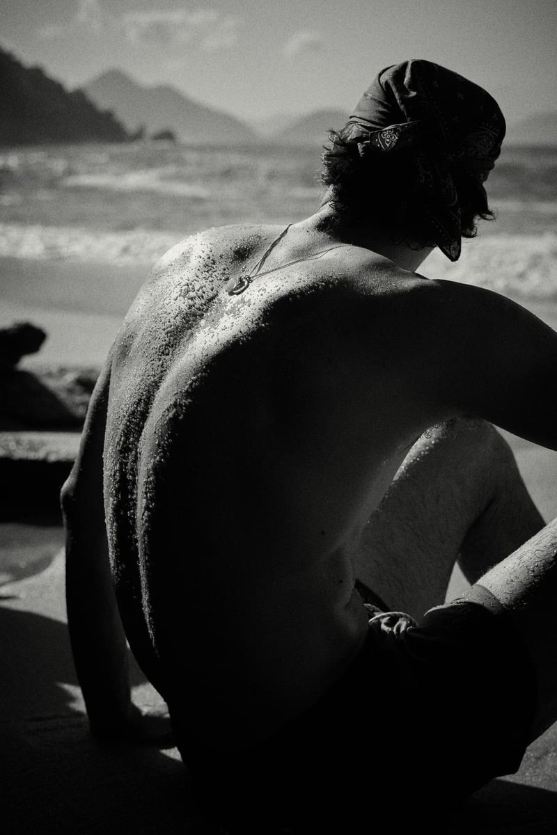 Francesco Ruggiero sits shirtless on the sand with his back to the camera, wearing a paisley bandana tied around his head and a thin pendant chain, salt and sand clinging to his shoulders as ocean waves and distant mountains stretch behind him in black and white