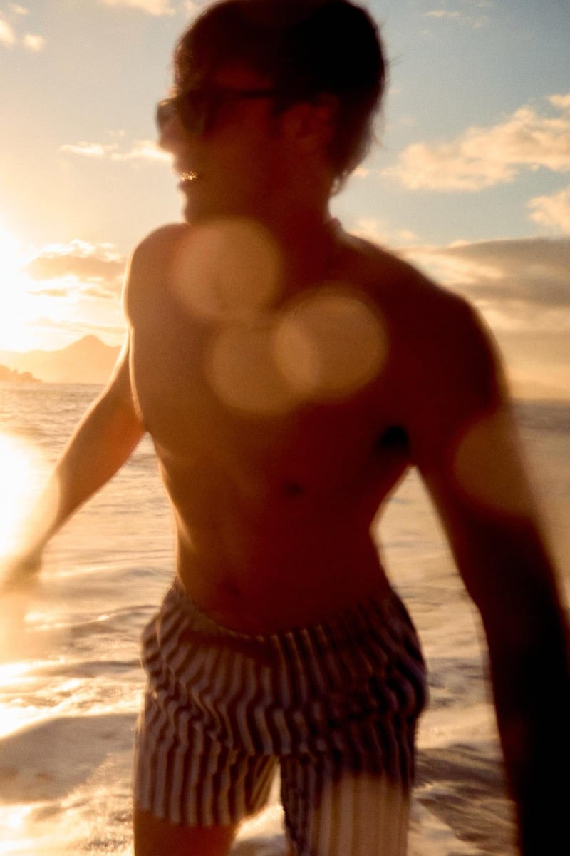 Francesco Ruggiero strides out of the surf at golden hour in navy and white vertical striped swim trunks and dark sunglasses, sunlight flaring across his bare chest