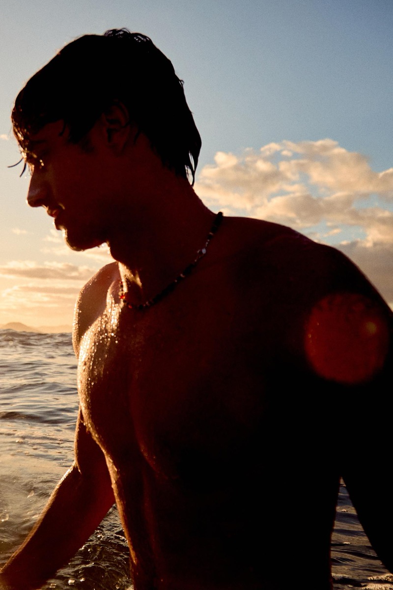 Francesco Ruggiero stands waist-deep in the ocean at sunset with wet hair pushed back, a multicolored beaded necklace at his throat, his torso outlined against a pink and orange sky