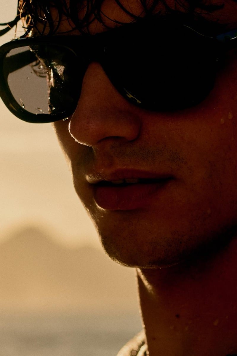 Tight close-up of Francesco Ruggiero in dark sunglasses, water droplets clinging to the lenses and stubble shading his jaw under golden afternoon light