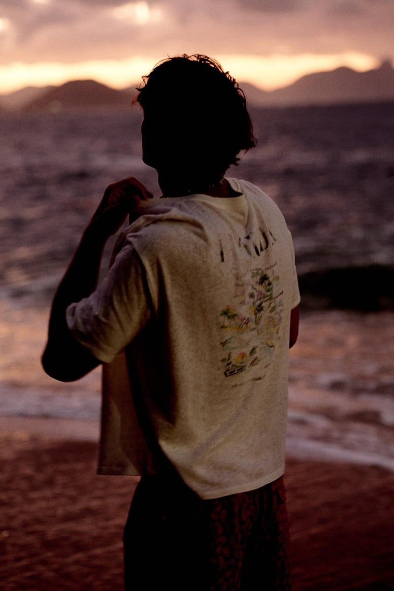 Francesco Ruggiero stands with his back to the camera at sunset, wearing a vintage-style graphic T-shirt with a tropical illustration and dark printed shorts, the violet ocean stretching to silhouetted mountains